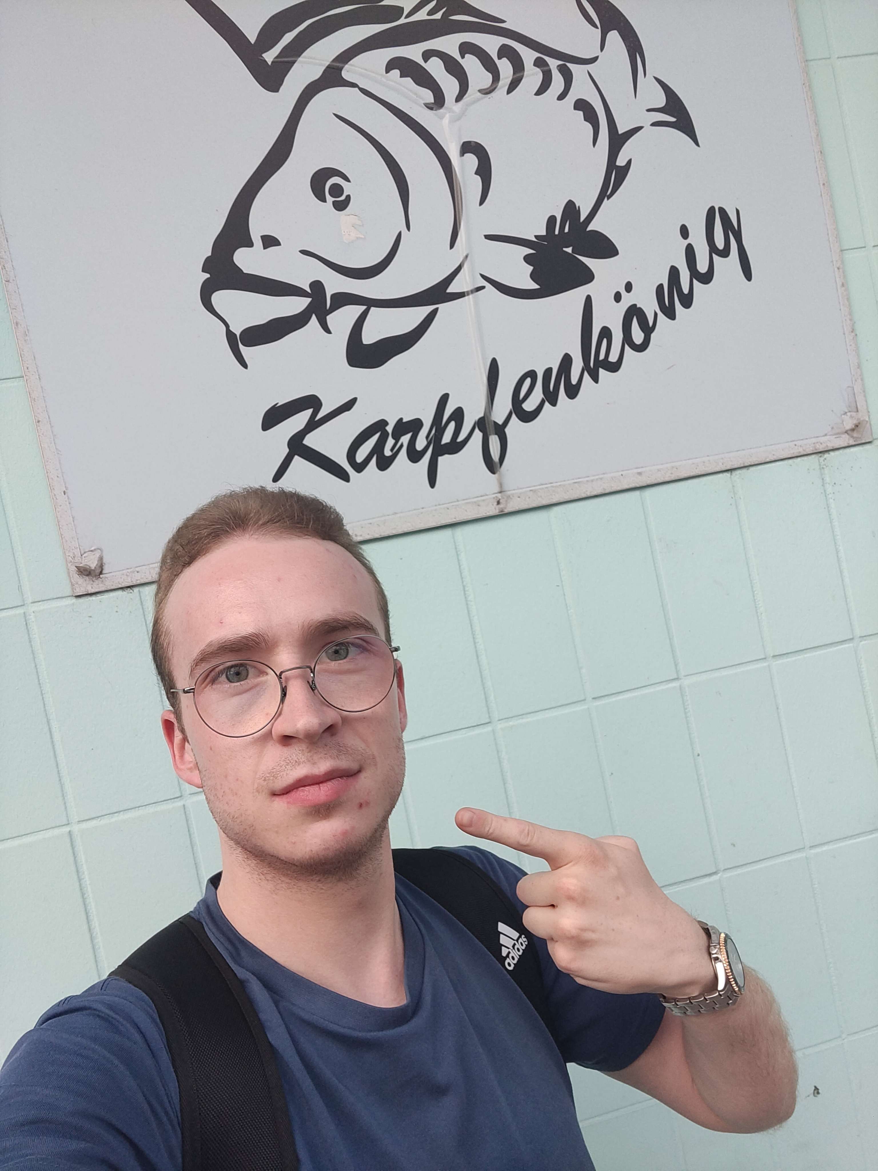 picture of a person standing in front of a sign saying capr king, pointing at himself, implying that he, indeed, is the carp king